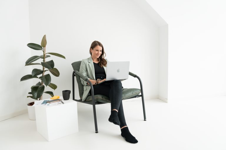 Es zeigt die Gründerin des Unternehmens, die auf einem Stuhl sitzt und mit dem Laptop auf dem Schoß arbeitet. Daneben ist eine Pflanze sowie ein kleiner Tisch mit Arbeitsmaterialien, Post-Its und einer Kaffeetasse. Das Bild ist sehr minimalistisch und klar.