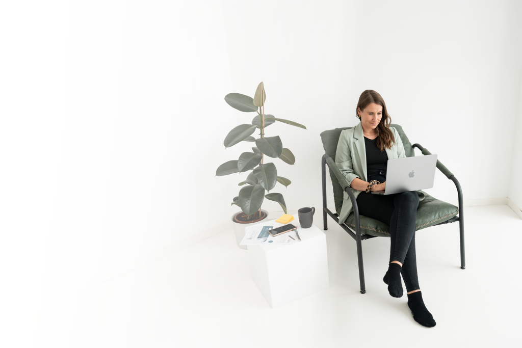 Es zeigt die Gründerin des Unternehmens, die auf einem Stuhl sitzt und mit dem Laptop auf dem Schoß arbeitet. Daneben ist eine Pflanze sowie ein kleiner Tisch mit Arbeitsmaterialien, Post-Its und einer Kaffeetasse. Das Bild ist sehr minimalistisch und klar.